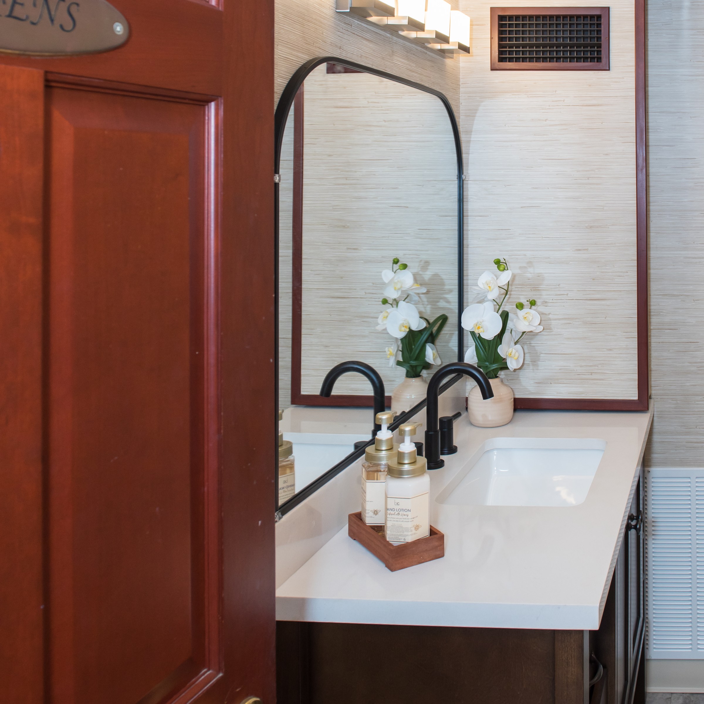 Elegant bathroom with flowers, black faucet, soap dispenser on white countertop, wooden door slightly open.