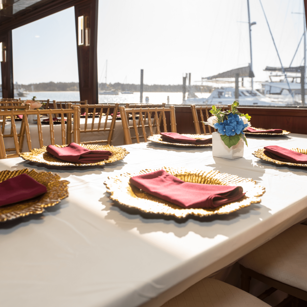 Elegant dining setup with gold plates, red napkins, and blue flowers, overlooking a marina view.