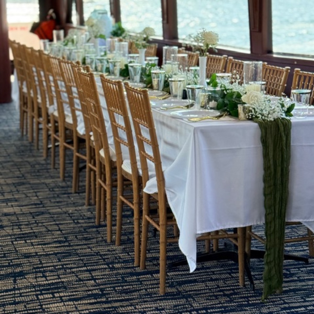 Long dining table with white cloth, flowers, chairs, by large windows with water view.