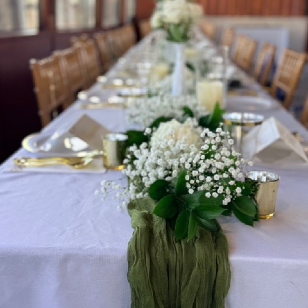 Elegant table setting with white flowers and green runner on white cloth, wooden chairs in background.