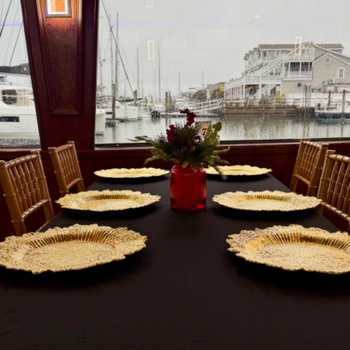 Table set with six ornate gold plates, red vase centerpiece, marina view through window.