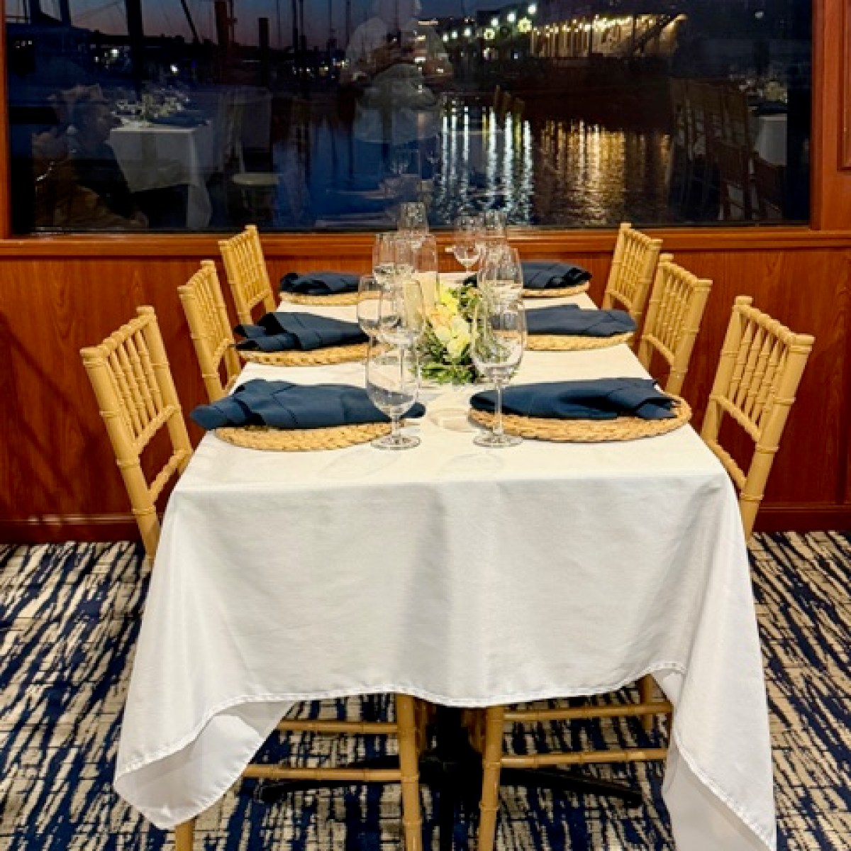 Table set for six with white cloth, gold chairs, and a marina view at sunset.