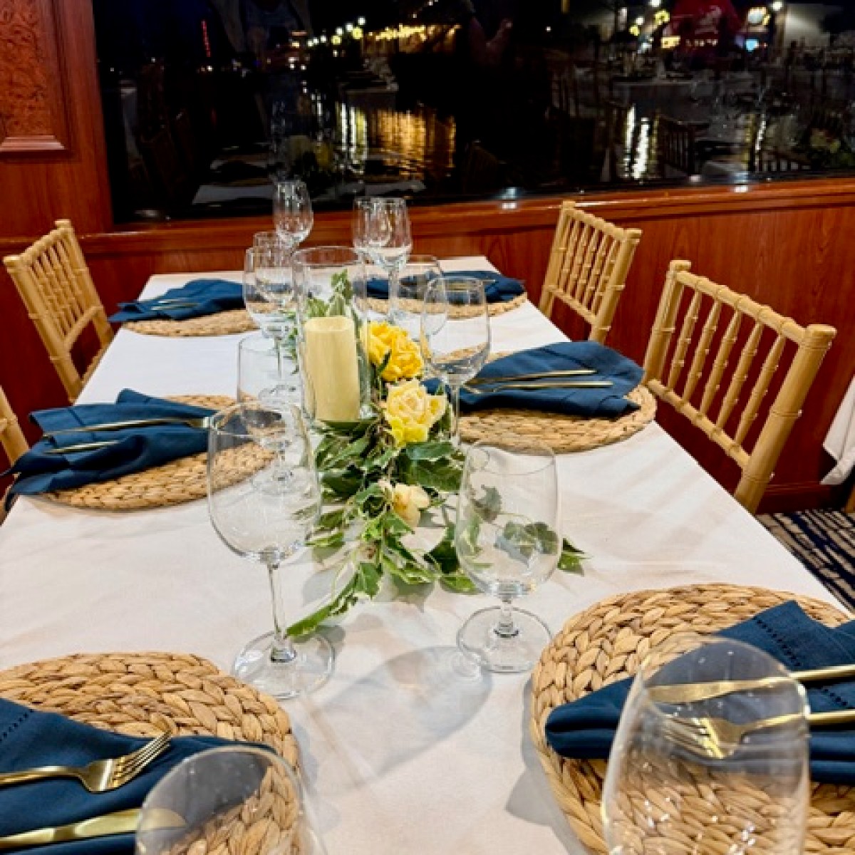 Elegant dining table set with yellow roses, placemats, and glassware, with city lights visible through window.