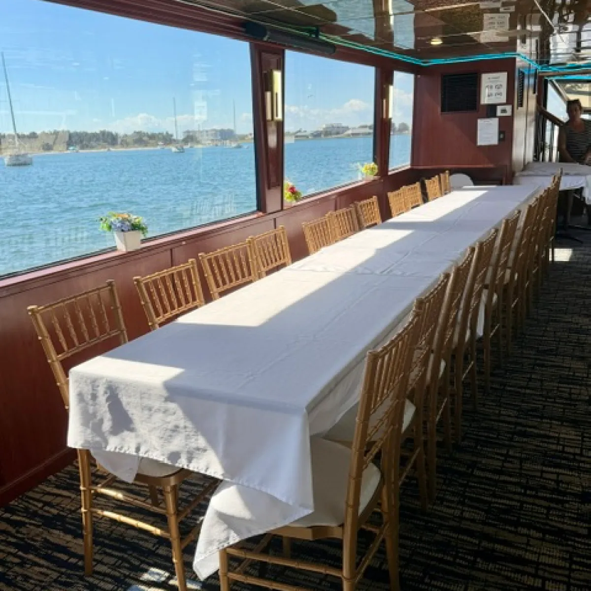 Long table with white cloth and chairs on a boat with ocean view through windows.