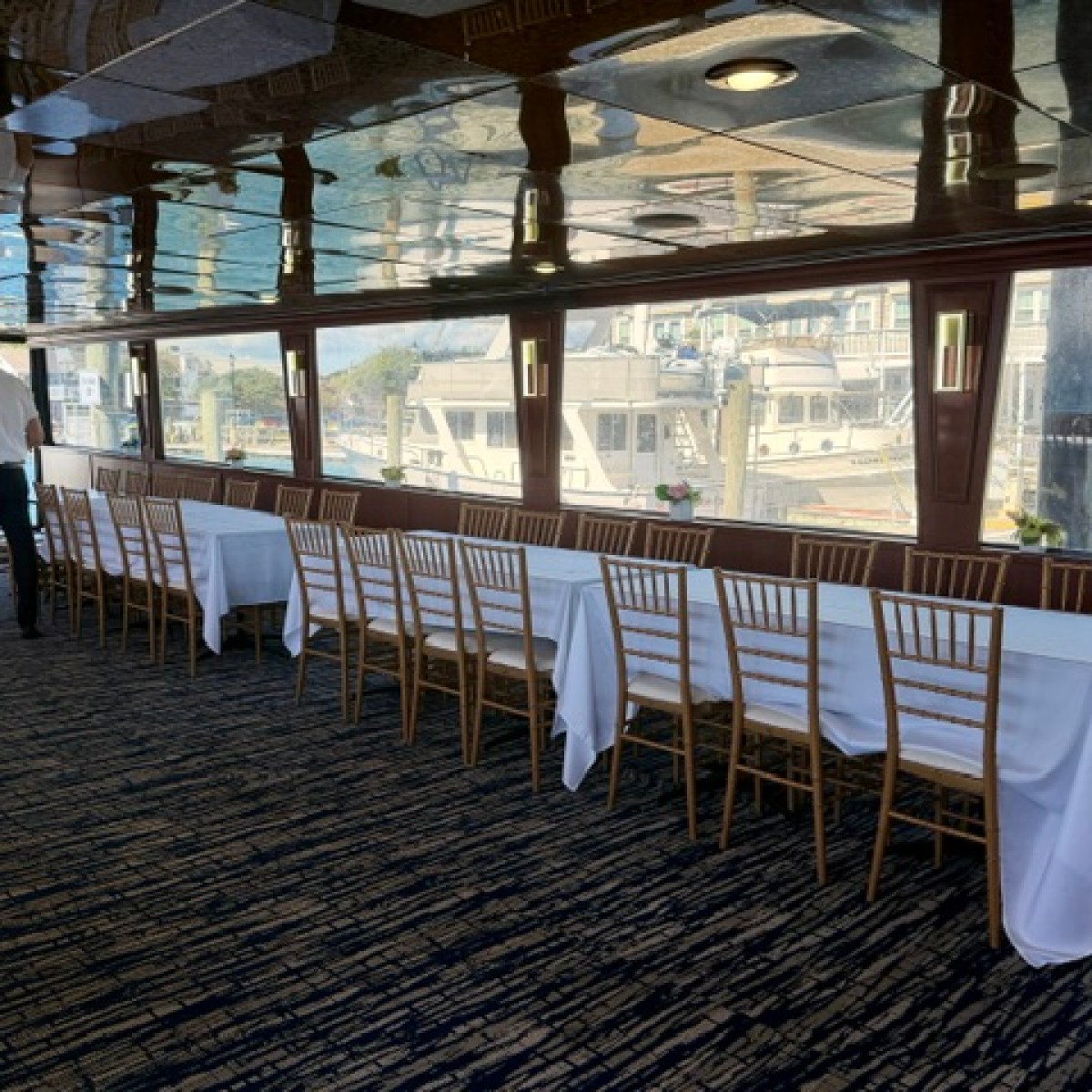 Empty dining area with long tables and a view of boats through large windows.