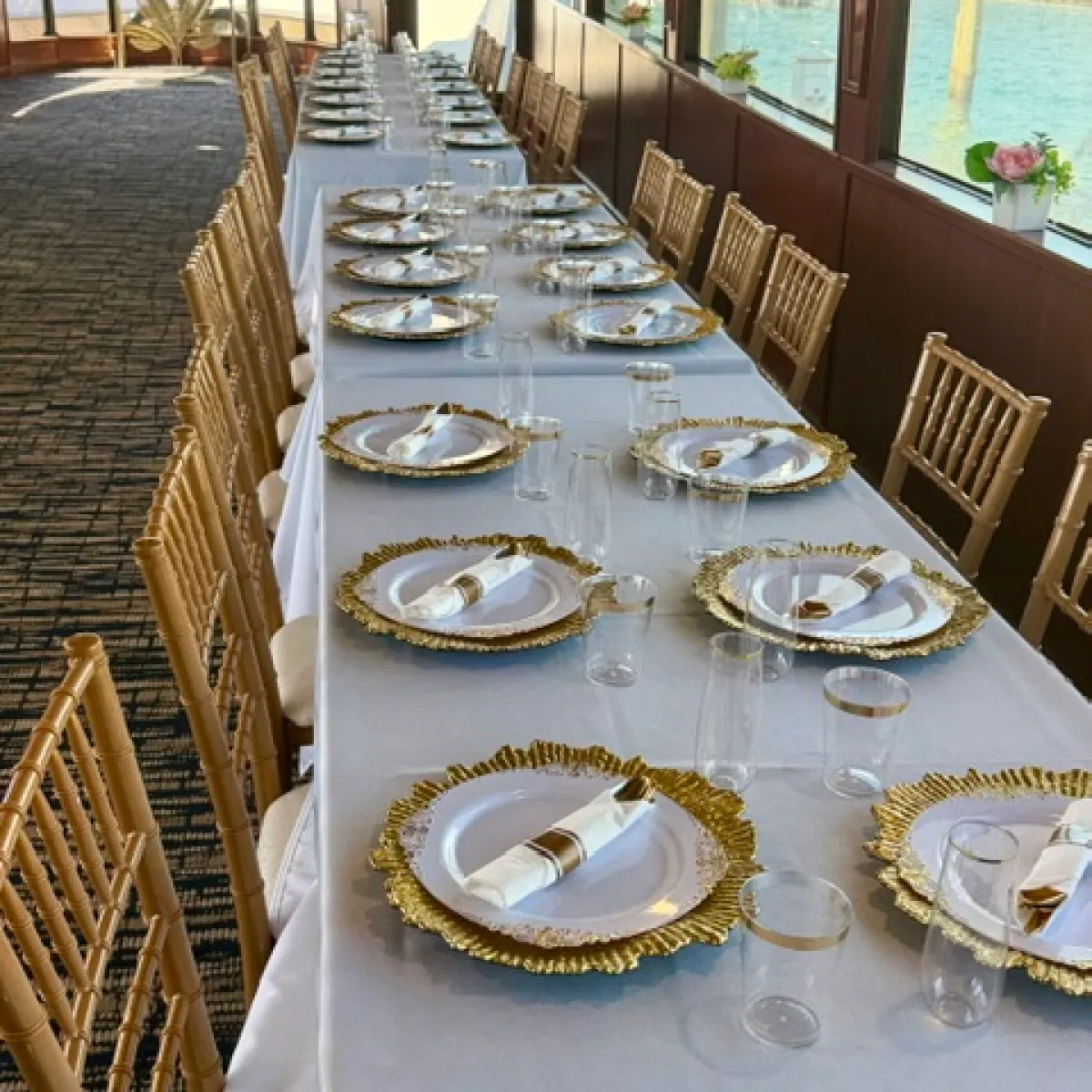 Long table set with white cloth, gold plates, and napkins by windows overlooking water.
