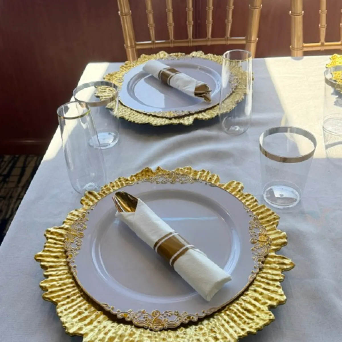 Elegant table setting with gold plates and napkin rings, clear glasses, and gold chairs.