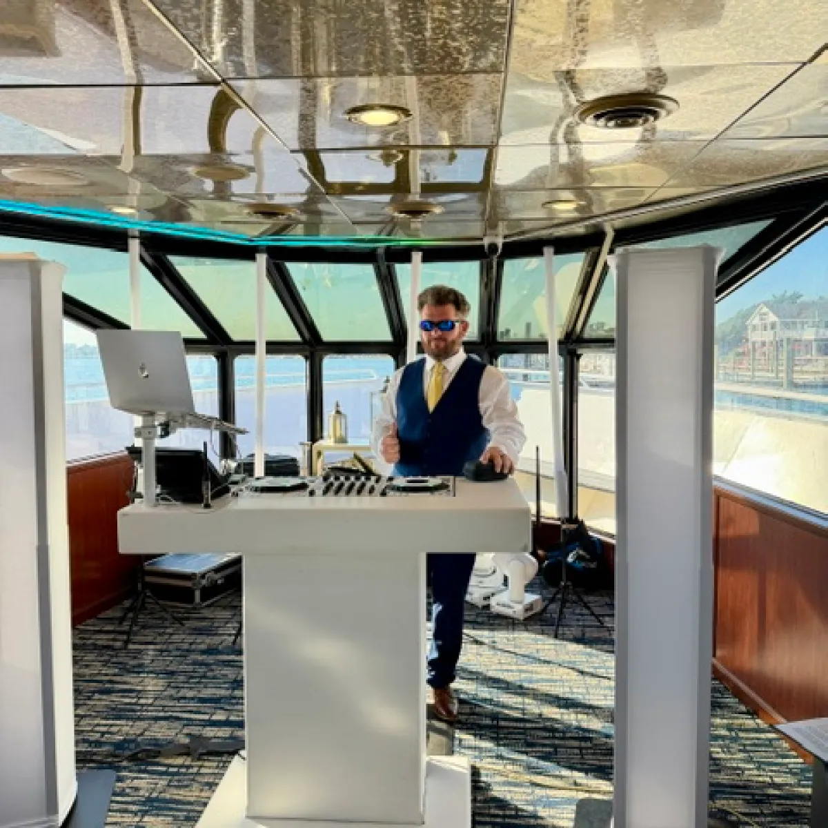 Person in suit with sunglasses at a DJ booth on a boat.